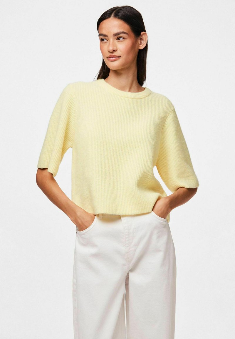 Żółty sweter dzianinowy w prążki z krótkimi rękawami i okrągłym dekoltem, w połączeniu z jasnymi spodniami o luźnym kroju i fakturze.