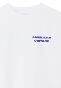 Valkoinen puuvillapaita, jossa on sininen "AMERICAN VINTAGE" -grafiikkateksti vasemmassa ylärintakappaleessa; lyhyet hihat ja pyöreä pääntie.