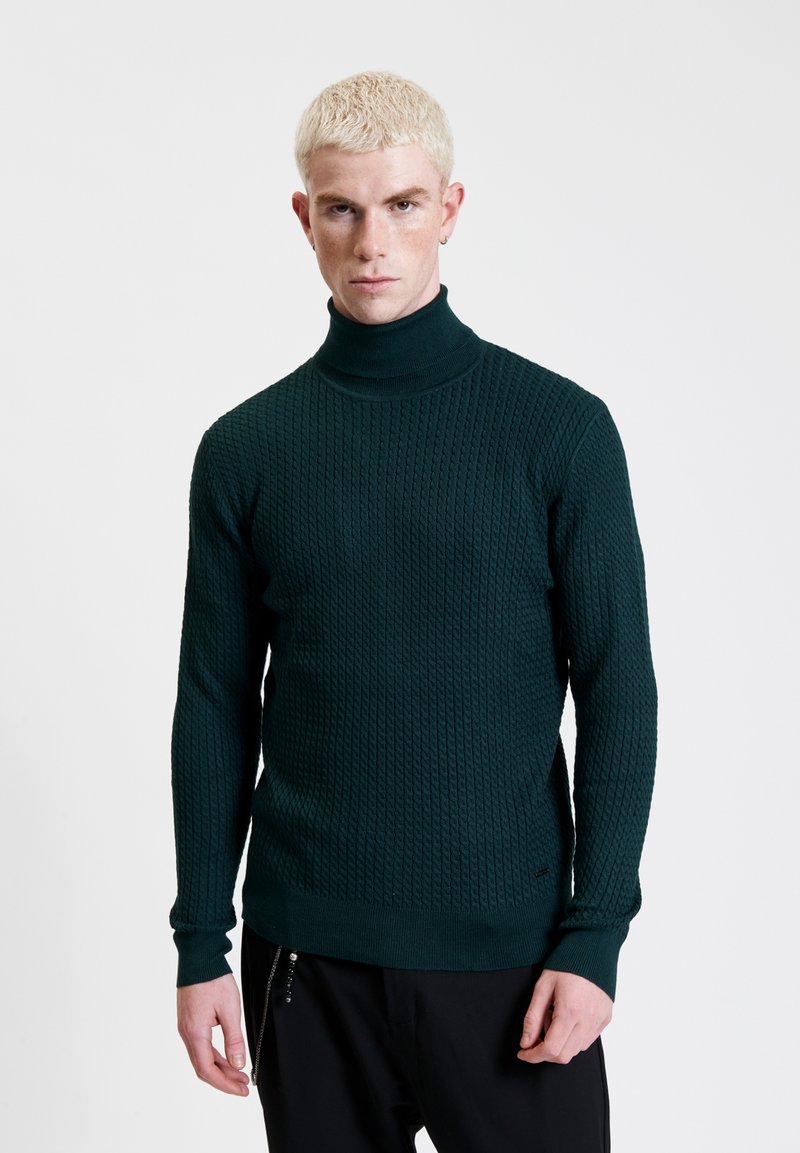 Maglione a collo alto in maglia verde scuro con motivo testurizzato, silhouette aderente e polsini a costine. Indossato con pantaloni neri.