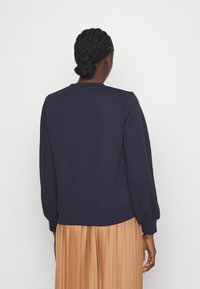 Maglione blu navy con maniche lunghe e polsini a coste. Presenta un collo rotondo e un orlo dritto, abbinato a una gonna beige plissettata.