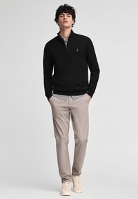 Ung man i svart ziptröja, rutmönstrad skjorta, beige byxor och beige sneakers står med händerna i fickorna mot en enkel bakgrund.