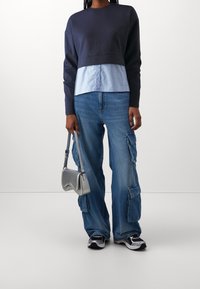Sweat-shirt court bleu marine superposé à une chemise à rayures bleu clair boutonnée, associé à un jean cargo bleu ample et un petit sac à main argenté.