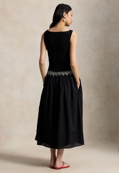 Pitkähiuksinen nainen, joka pitää käsiään taskuissa. Hänellä on yllään hihaton musta mekko, jossa on pitsinen vyötärön yksityiskohta, ja punaiset tasapohjaiset sandaalit.