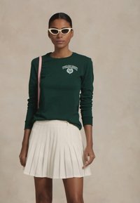 Zöld, hosszú ujjú ing Wimbledon logóval, fehér rakott szoknyával párosítva. A modellen fehér, macskatempójú napszemüveg is található.