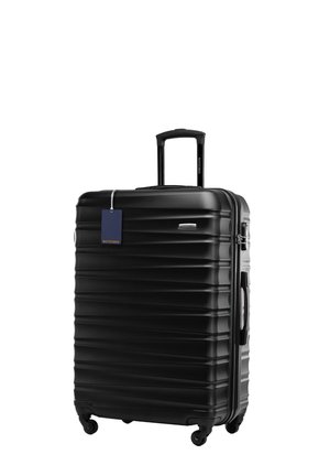 Valise rigide noire avec texture nervurée, poignée extensible, quatre roues et une étiquette à bagages bleue. Forme rectangulaire compacte.