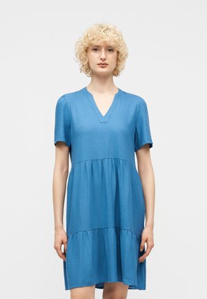 Persona rubia con cabello rizado corto, vestida con un vestido azul de manga corta y capas, con escote en V, de pie frente a un fondo claro y liso.