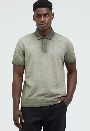Khaki green polo shirt made of textured fabric, featuring a collar, two-button placket, and short sleeves, paired with dark pants.