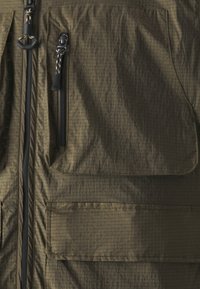 Olīvu zaļa veste ar strukturētu, vieglu audumu. Aprīkota ar divām melnām rāvējslēdzēja kabatām un lielu priekšējo kabatu ar klapām. Izturīga šuvuma izpilde.