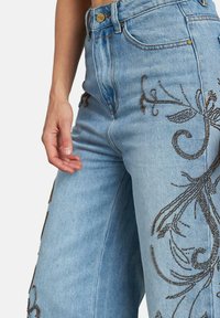Jeans de mezclilla azul de cintura alta con intrincados bordados de cuentas marrones en patrones florales. Presentan bolsillos frontales y detalle de botón metálico.