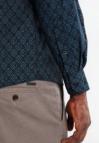 Donkerblauwe lange mouwen overhemd met geometrische patronen; gecombineerd met lichtbruine broeken; heeft knoopmanchetten en een zakdetail.