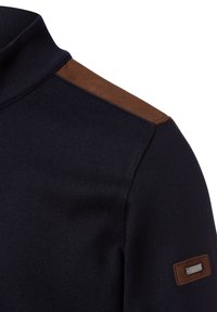 Maglione blu navy con colletto alto, dotato di dettagli in suede marrone sulle spalle e una piccola patch logo argentata sulla manica. Texture lavorata a maglia.