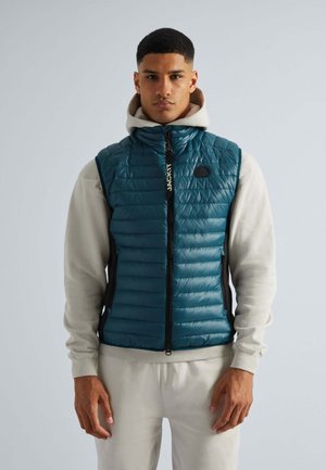 Teal puffer gilet met horizontale stiksels, zwart ritssluiting en logo-detail, gedragen over een lichtgrijze hoodie, gecombineerd met grijze sweatpants.
