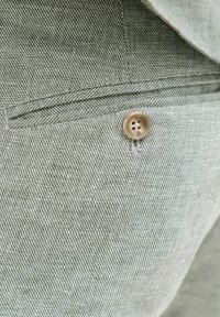 Κοντινή λήψη μπεζ υφασμάτινου παντελονιού με υφή, με καφέ κουμπί τετράγωνης τρύπας και οριζόντια τσέπη με φάσα πάνω από το κουμπί.