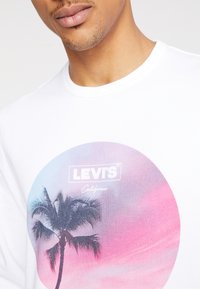 T-shirt en coton blanc avec un graphisme circulaire dégradé rose et bleu, silhouette de palmier et texte "LEVI'S California".
