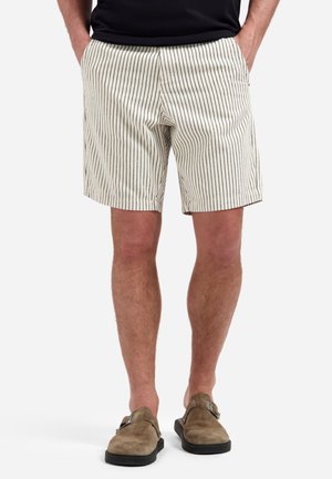 Man draagt beige korte broek met zwarte verticale strepen, zwart shirt en bruine suède instapschoenen, staat met handen in de zakken.