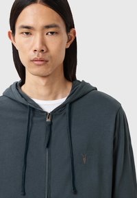 Felpa con zip color verde scuro, dotata di cordino, caratterizzata da un logo discreto sul petto e un tessuto morbido e strutturato.