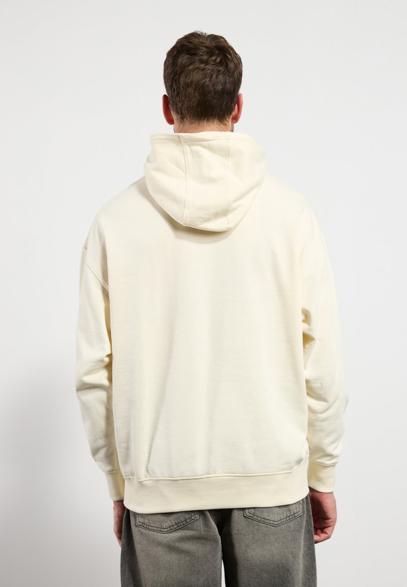 Sweat-shirt à capuche de couleur crème, au texture douce, coupe décontractée et avec des poignets et un ourlet côtelés. Présente un design simple et sans embellissement.