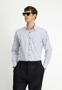 Camicia bianca con righe verticali blu, colletto button-down e una tasca sul petto, abbinata a pantaloni scuri sartoriali e occhiali da sole neri.