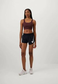 Sujetador deportivo de color burdeos, combinado con pantalones cortos negros que presentan un logo blanco. El conjunto se complementa con zapatillas deportivas en un suave tono rosa.