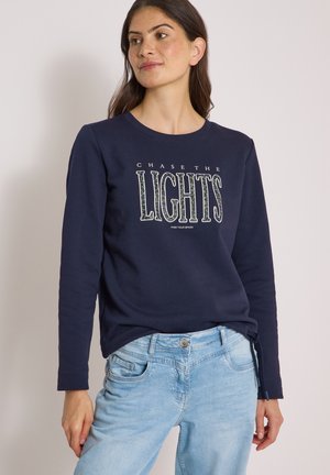 Frau trägt einen dunkelblauen Pullover mit dem Text "CHASE THE LIGHTS FIND YOUR SPARK" und hellblaue Jeans und steht vor einem einfarbigen Hintergrund.