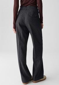 Femme portant un pantalon large taille haute gris foncé avec une pochette bouton au dos et un haut à manches longues côtelé marron, se tenant sur fond blanc.