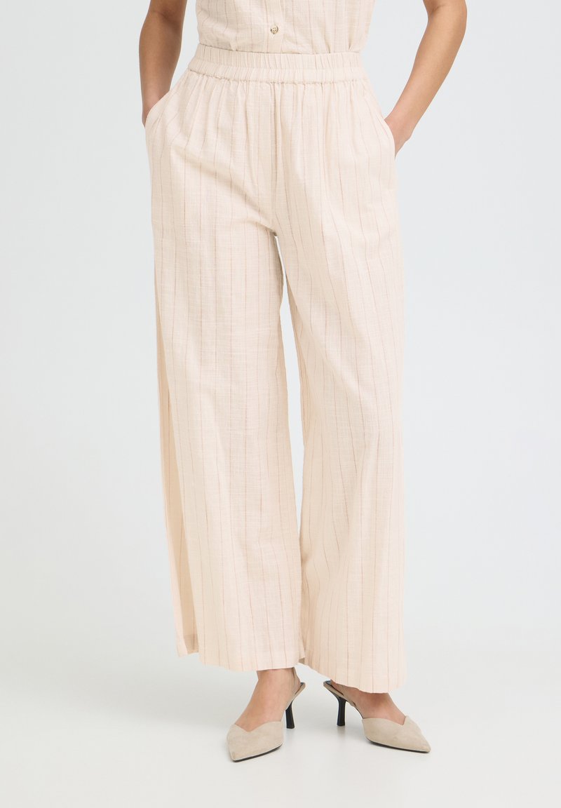 Femme portant un pantalon large beige à fines rayures verticales et des mules à talons pointues beige sur fond blanc.