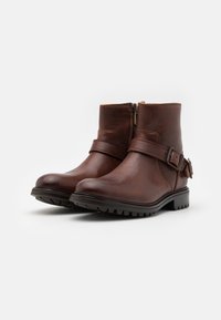 Botas de tobillo de cuero marrón con un acabado suave, cremallera lateral y acentos de cinturón ajustable. Cuenta con una suela de goma texturizada para mayor agarre.