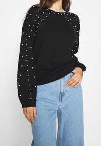 Maglione nero lavorato a maglia con decorazioni di perle sulle maniche, scollatura rotonda e polsini a costine. Abbinato a jeans a gamba larga blu chiaro.