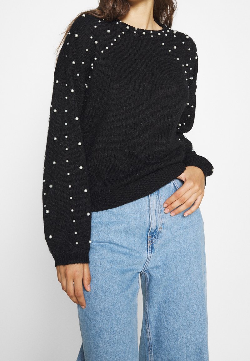 Maglione nero lavorato a maglia con decorazioni di perle sulle maniche, scollatura rotonda e polsini a costine. Abbinato a jeans a gamba larga blu chiaro.