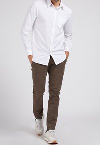 Wit overhemd met lange mouwen en knopen, voorzien van een zak, gecombineerd met olijfgroene chino's en witte sneakers met beige accenten.