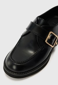 Black leather shoe featuring a smooth finish, rounded toe, adjustable strap with a gold buckle, and decorative stitching along the sole.