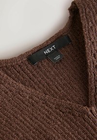 Pull en maille marron texturé avec un col en V et une étiquette en tissu noire indiquant "NEXT" et "Made in China".