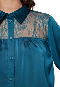 Tealblus med satinkänsla och spetsdetalj vid axlarna. Har en klassisk krage, knäppning med knappar och mjuka veck under spetssektionen.