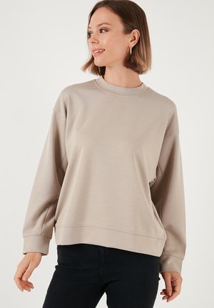 Beež ümmarguse kaelusega sviiter pikade varrukatega, pehme tekstuuriga ja lõdva lõikega; koos tumedate teksadega. Minimalistlik tikand detail hemil.