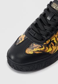 Sapatilha preta com um upper em pele texturizada, um vibrante design floral amarelo e laranja, e uma sola de borracha embutida com "JEANS COUTURE."