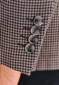 Brązowa i niebieska tkanina w wzór houndstooth z czterema przezroczystymi guzikami oraz z detalicznym przeszyciem na dopasowanym rękawie.