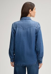 Camicia di jeans blu scuro con maniche lunghe, polsini con bottoni e colletto classico. Presenta un dettaglio di yoke nella parte posteriore e un orlo curvo.