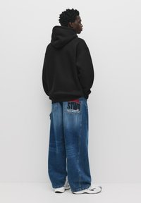 Black hoodie with a relaxed fit, paired with oversized light blue denim jeans featuring a red and black graphic patch on the back.