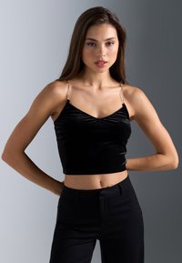 Top corto de terciopelo negro con escote en V y tirantes con detalles de cadena, exhibido sobre un suave fondo gris. Textura suave, diseño ajustado.