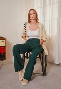 Junge Frau mit roten Haaren sitzt im Rollstuhl und trägt einen beigen Cardigan, ein weißes Oberteil, grüne Hose und beige Slipper in Innenräumen.
