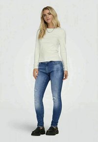 White long-sleeve shirt and blue skinny jeans with distressed details. Black footwear with a thick sole, standing against a plain background.