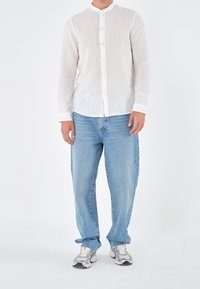 Chemise en lin blanc avec col mao, boutonnée sur le devant, associée à un jean bleu clair et des baskets sportives argentées. Fond uni.