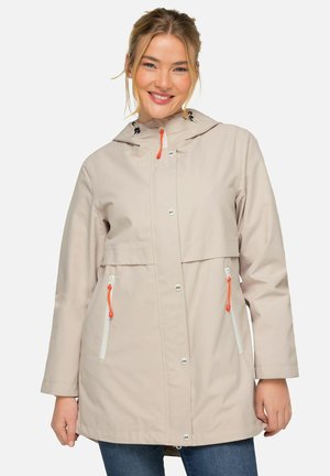 Beige wasserfeste Jacke mit Kapuze, ausgestattet mit orangefarbenen Kordeln, Druckknöpfen und zwei Reißverschlusstaschen mit weißen Akzenten.