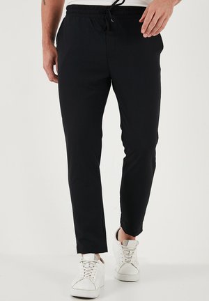 Sorte atletiske bukser med snøre i taljen, slim fit, elastisk stof og sidelommer, parret med hvide sneakers. Glat tekstur.