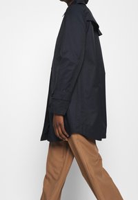 Persona caminando con un abrigo azul marino oscuro y pantalones de pierna ancha marrón claro, frente a un fondo claro y liso.