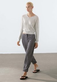 Lichtgrijze, lange mouwen V-hals trui gecombineerd met een op maat gemaakte, donkergrijze broek met plooien. Zwarte teenslippers maken de look compleet.