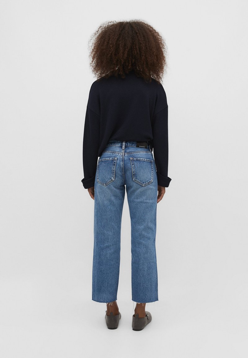 Persona con cabello rizado vistiendo un suéter negro, jeans rectos azules y zapatos grises tipo slip-on, de pie frente a un fondo blanco liso.