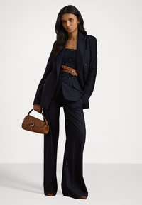 Marineblauw gestreept pak met wijde broekspijpen en een op maat gemaakte blazer. Een bruine leren riem en handtas accentueren het professionele ontwerp van de outfit.