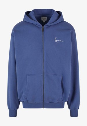 Zilā apmava ar rāvējslēdzēju, izgatavota no mīkstas auduma, ar priekšējiem kabatām, ribotām aprocēm un elastīgu jostasvietu. Uz krūtīm atrodas mazs balts logo.