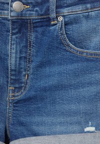 Gros plan sur un jean en denim bleu montrant une poche avant, une boucle de ceinture, un bouton en métal, une couture jaune et une petite zone usée près de l'ourlet.